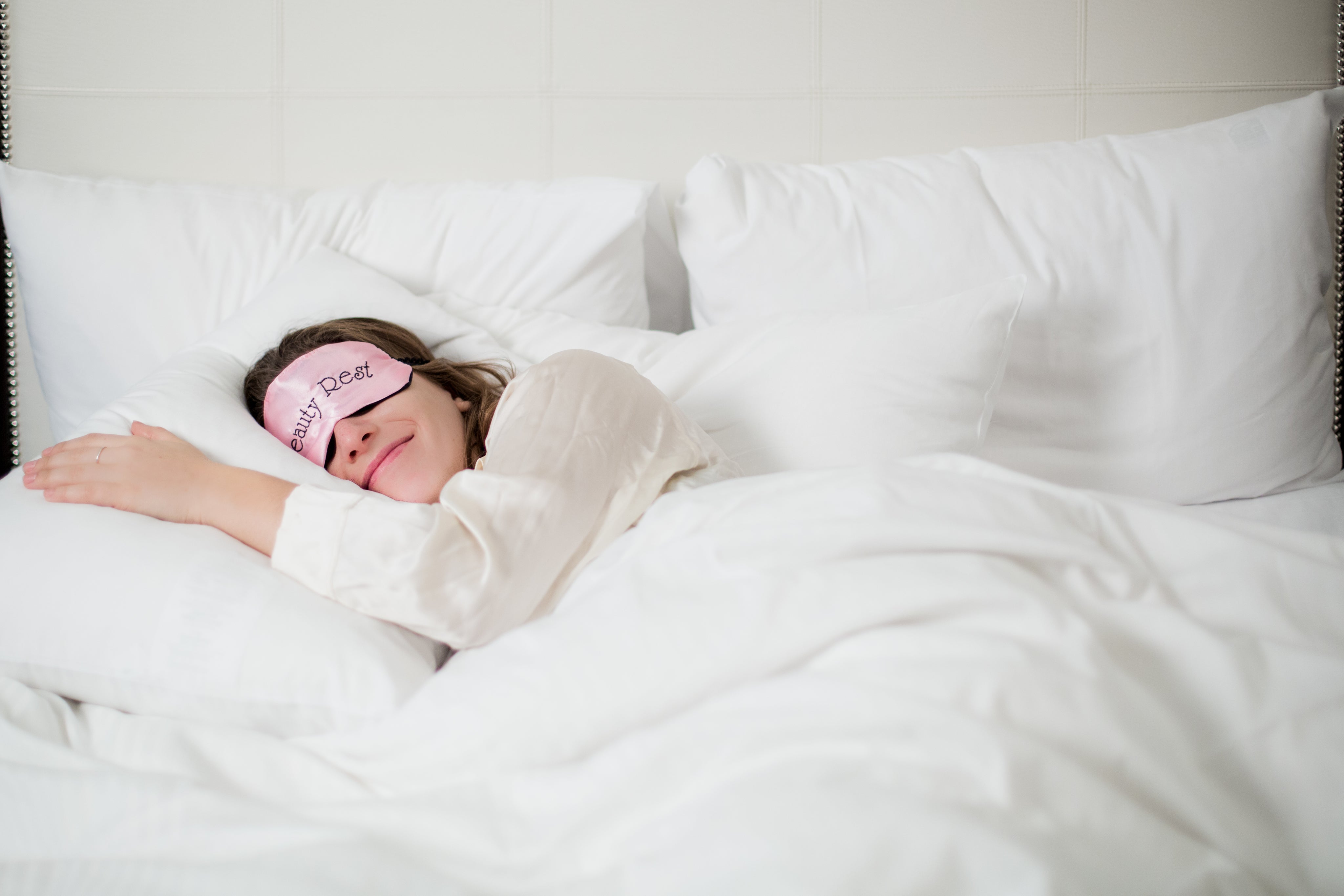 Smiling woman peacefully sleeping on a bed with white bedsheets and pillowcases, wearing a pink sleep mask