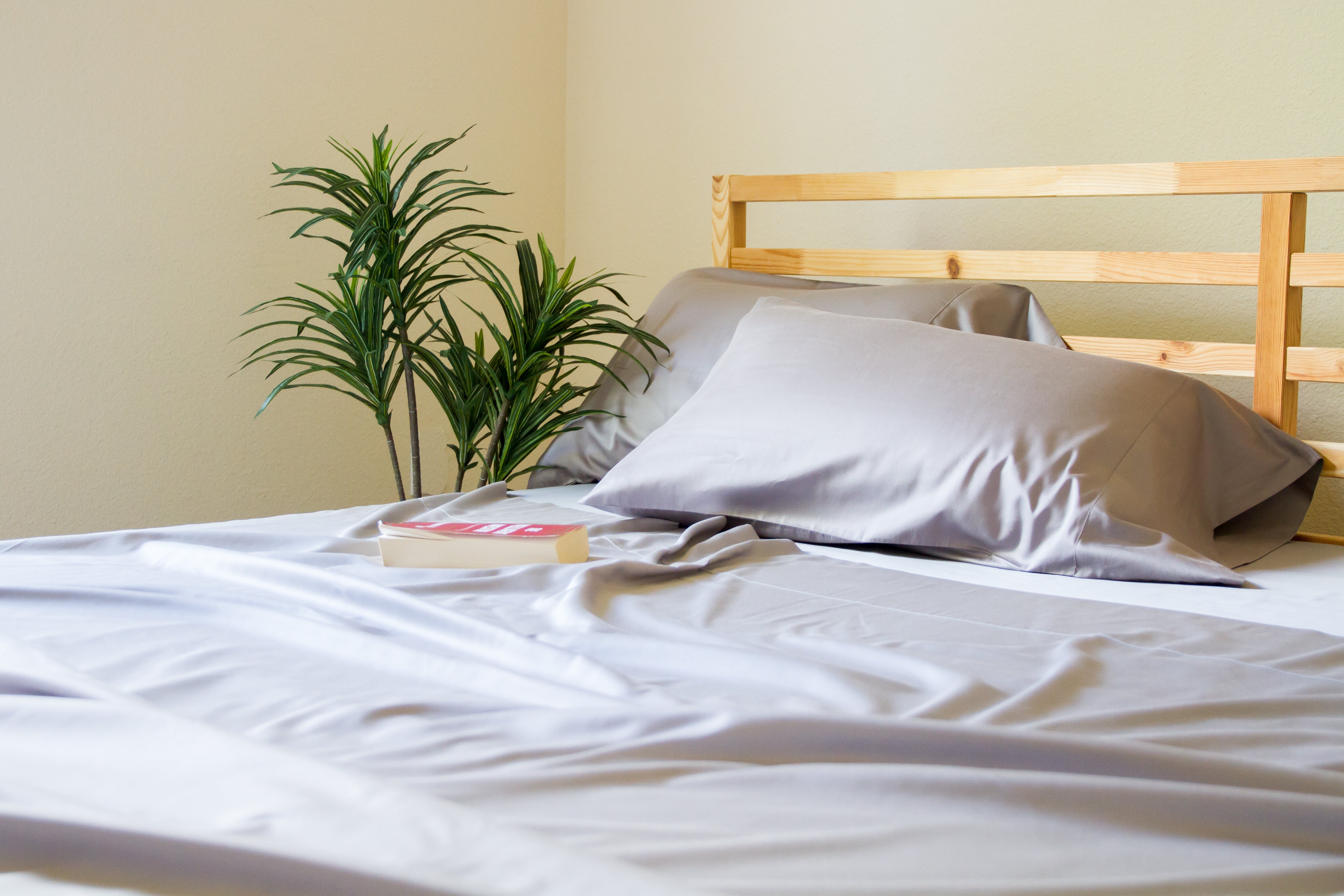 Bed with Gray sheets, wooden headboard, and a plant in the corner.