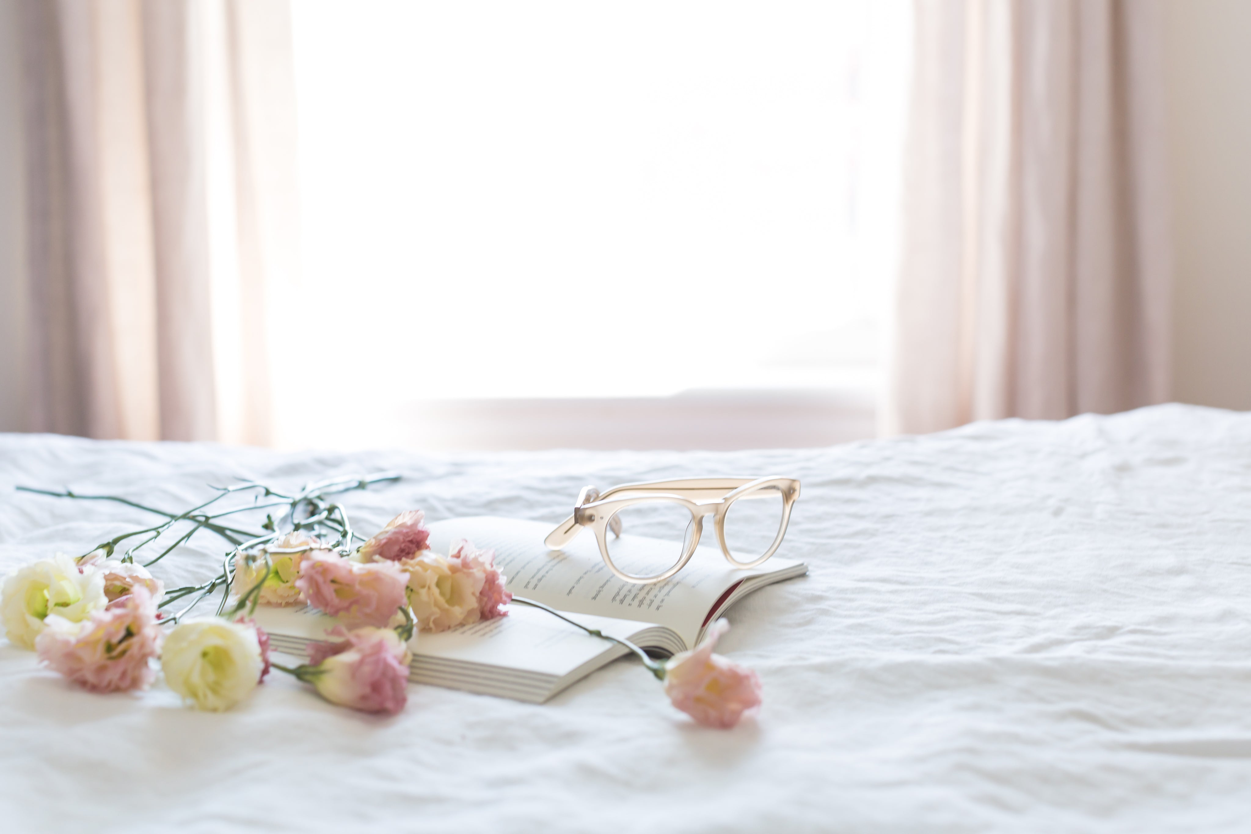 Spectacles and flowers resting on an open book placed on a bed with a white bedsheet