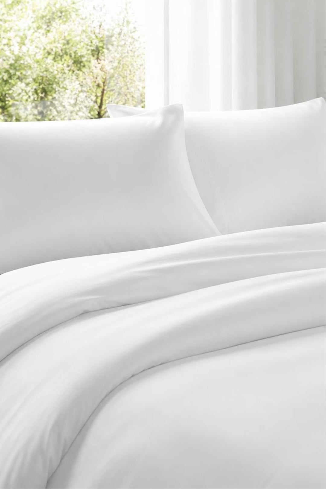 White bedding set with two pillows and a duvet cover, styled on a bed in front of a bright window with soft light and blurred greenery outside.